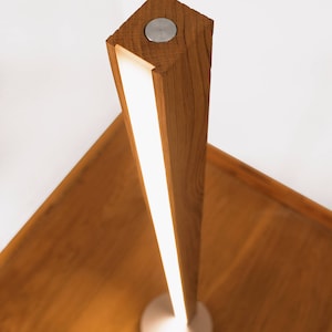 May include: A modern wooden table lamp with a rectangular, light-emitting column. The lamp has a brushed metal base and a small, circular metal button on top. The wood has a warm, natural tone, and the light is a soft white.