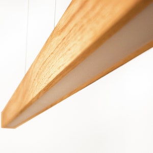May include: A modern, rectangular wooden light fixture with a warm, natural wood grain finish. The light fixture has a recessed, white, illuminated panel. The fixture is suspended from the ceiling by thin wires, creating a minimalist aesthetic.