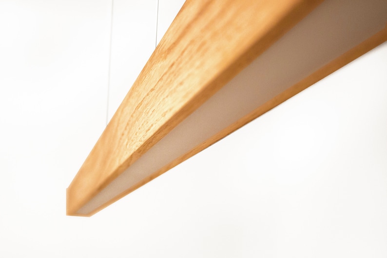 May include: A modern, rectangular wooden light fixture with a warm, natural wood grain finish. The light fixture has a recessed, white, illuminated panel. The fixture is suspended from the ceiling by thin wires, creating a minimalist aesthetic.