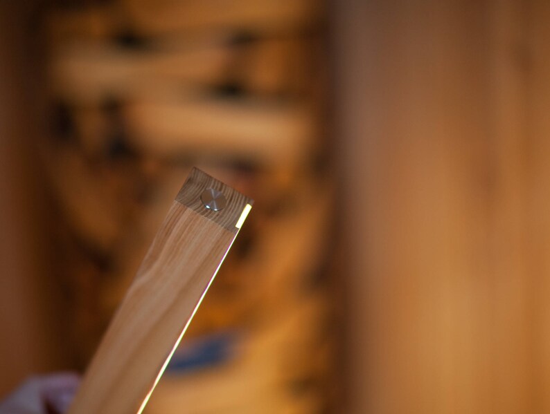 K&ouml;nnte beinhalten: Nahaufnahme eines Holzbretts mit hellfarbiger, nat&uuml;rlicher Holzmaserung. Das Ende des Brettes hat eine kleine Metallbefestigung. Das Holz wird von hellem Licht beleuchtet, was ein warmes, einladendes Gef&uuml;hl erzeugt.