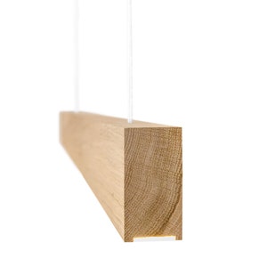 Puede incluir: Una lámpara rectangular de madera suspendida por dos cables blancos. La lámpara está hecha de madera de color claro, con vetas visibles y una fuente de luz empotrada en la parte inferior. La lámpara se muestra sobre un fondo blanco.