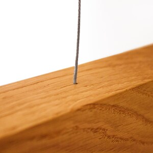 May include: Close-up of a wooden beam with a thin, gray wire extending from a small hole. The wood has a warm, reddish-brown tone and visible grain. The wire appears to be made of a twisted metal.