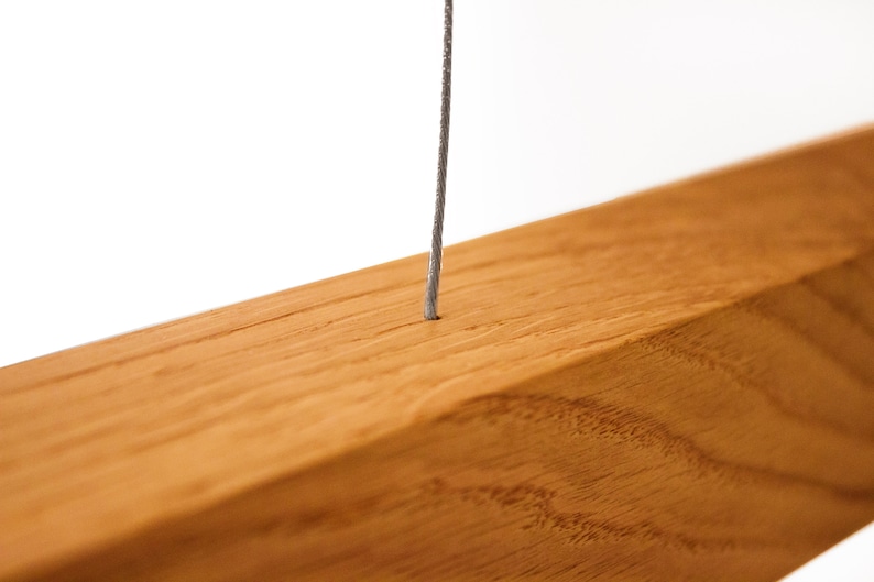 May include: Close-up of a wooden beam with a thin, gray wire extending from a small hole. The wood has a warm, reddish-brown tone and visible grain. The wire appears to be made of a twisted metal.