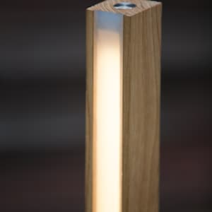 K&ouml;nnte beinhalten: Eine rechteckige Holzleuchte mit einem warmen, leuchtenden Lichtstreifen in der Mitte. Die Leuchte besteht aus hellfarbenem Holz und hat eine quadratische Oberseite mit einem kleinen, kreisf&ouml;rmigen Metallakzent.
