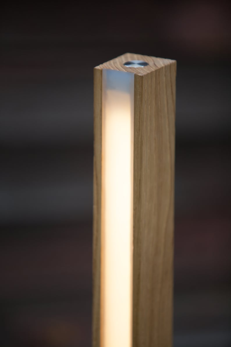 K&ouml;nnte beinhalten: Eine rechteckige Holzleuchte mit einem warmen, leuchtenden Lichtstreifen in der Mitte. Die Leuchte besteht aus hellfarbenem Holz und hat eine quadratische Oberseite mit einem kleinen, kreisf&ouml;rmigen Metallakzent.