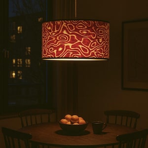 May include: A pendant lamp with a cylindrical shade featuring a red and cream abstract pattern hangs above a round wooden dining table. A bowl of oranges and a mug sit on the table, surrounded by chairs.