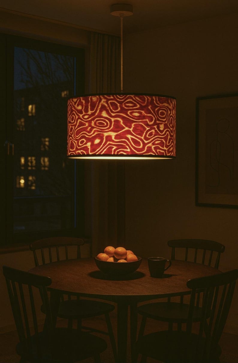 May include: A pendant lamp with a cylindrical shade featuring a red and cream abstract pattern hangs above a round wooden dining table. A bowl of oranges and a mug sit on the table, surrounded by chairs.