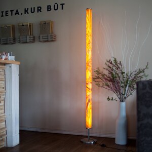 May include: A tall, illuminated floor lamp with a marbled orange and yellow pattern. The lamp stands on a silver base. The wall behind the lamp has the text "VIETA, KUR BŪT" in black.