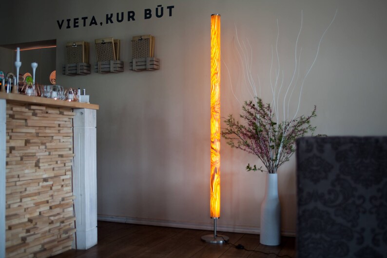 May include: A tall, illuminated floor lamp with a marbled orange and yellow pattern. The lamp stands on a silver base. The wall behind the lamp has the text "VIETA, KUR BŪT" in black.