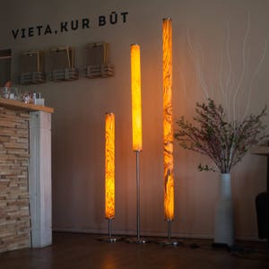 May include: Three illuminated floor lamps with a warm, amber glow. The lamps have a cylindrical shape with a wood grain pattern. The lamps are of varying heights, standing on silver metal bases. The background includes a wall with the text "VIETA.KUR B&Uuml;T".
