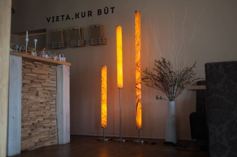 May include: Three illuminated floor lamps with a warm, amber glow. The lamps have a cylindrical shape with a wood grain pattern. The lamps are of varying heights, standing on silver metal bases. The background includes a wall with the text "VIETA.KUR B&Uuml;T".