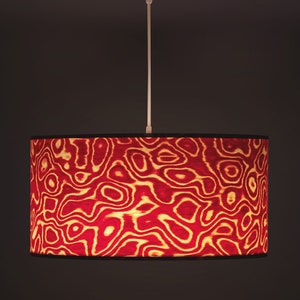 May include: A pendant light fixture with a cylindrical lampshade. The lampshade features a red and gold abstract pattern. The light fixture has a white ceiling mount and a white cord. The background is a dark brown.