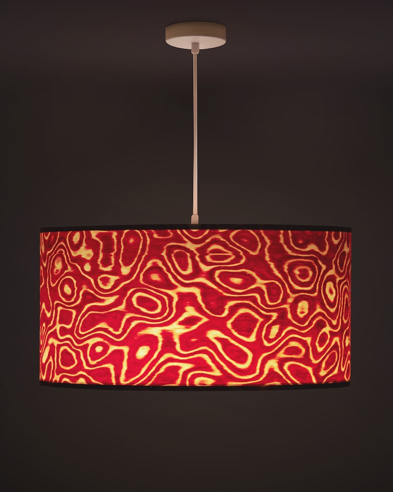 May include: A pendant light fixture with a cylindrical lampshade. The lampshade features a red and gold abstract pattern. The light fixture has a white ceiling mount and a white cord. The background is a dark brown.