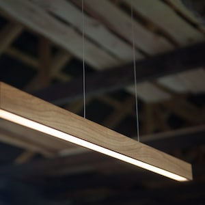 May include: A rectangular wooden pendant light fixture with a warm, natural wood grain finish. The light is suspended by thin wires and emits a soft, diffused glow. The fixture is approximately 48 inches long.