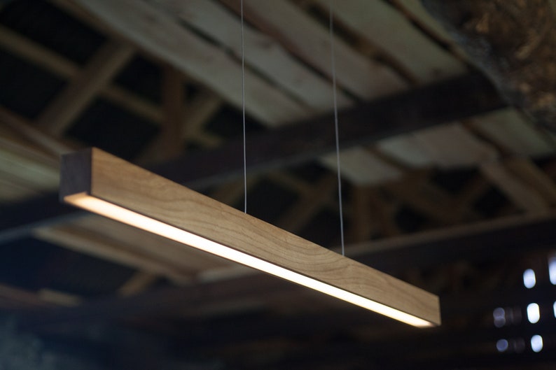 May include: A rectangular wooden pendant light fixture with a warm, natural wood grain finish. The light is suspended by thin wires and emits a soft, diffused glow. The fixture is approximately 48 inches long.