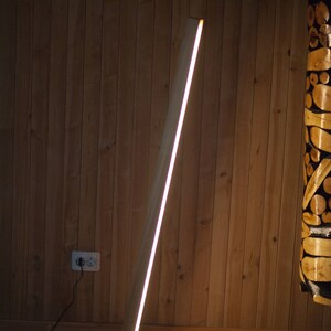 K&ouml;nnte beinhalten: Eine moderne Stehlampe aus Holz mit einer hellen, linearen Lichtquelle. Die Lampe hat eine helle Holzoberfl&auml;che und einen kreisf&ouml;rmigen Sockel. Das Licht hat eine warme wei&szlig;e Farbe und die Lampe steht vor einer Holzwand.