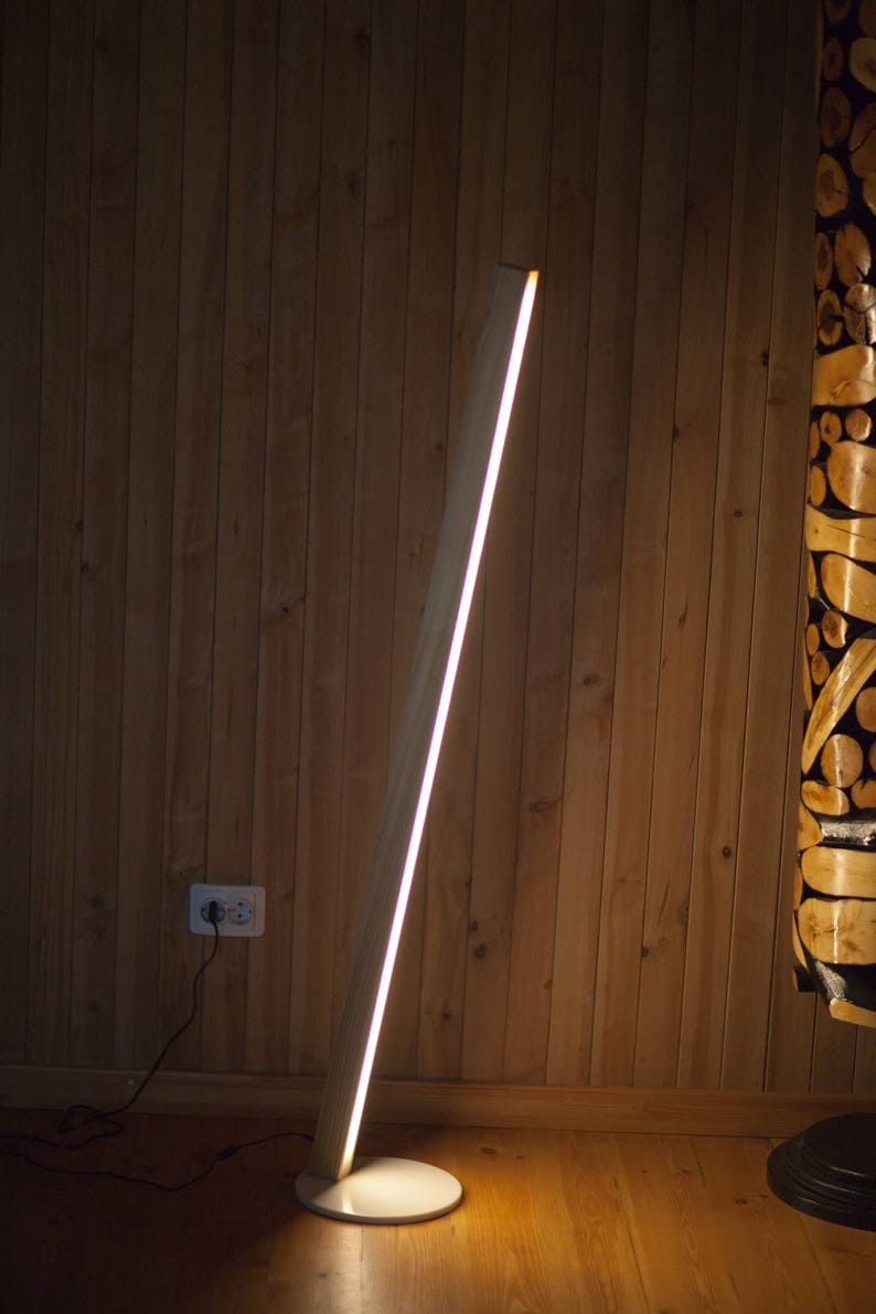 K&ouml;nnte beinhalten: Eine moderne Stehlampe aus Holz mit einer hellen, linearen Lichtquelle. Die Lampe hat eine helle Holzoberfl&auml;che und einen kreisf&ouml;rmigen Sockel. Das Licht hat eine warme wei&szlig;e Farbe und die Lampe steht vor einer Holzwand.