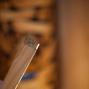 K&ouml;nnte beinhalten: Nahaufnahme eines Holzbretts mit hellfarbiger, nat&uuml;rlicher Holzmaserung. Das Ende des Brettes hat eine kleine Metallbefestigung. Das Holz wird von hellem Licht beleuchtet, was ein warmes, einladendes Gef&uuml;hl erzeugt.