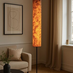 May include: A tall, cylindrical floor lamp with a warm, wood-grain patterned shade. The shade is illuminated, casting a soft glow. The lamp has a slender, black metal stand. The lamp is in a living room setting with a sofa and artwork.