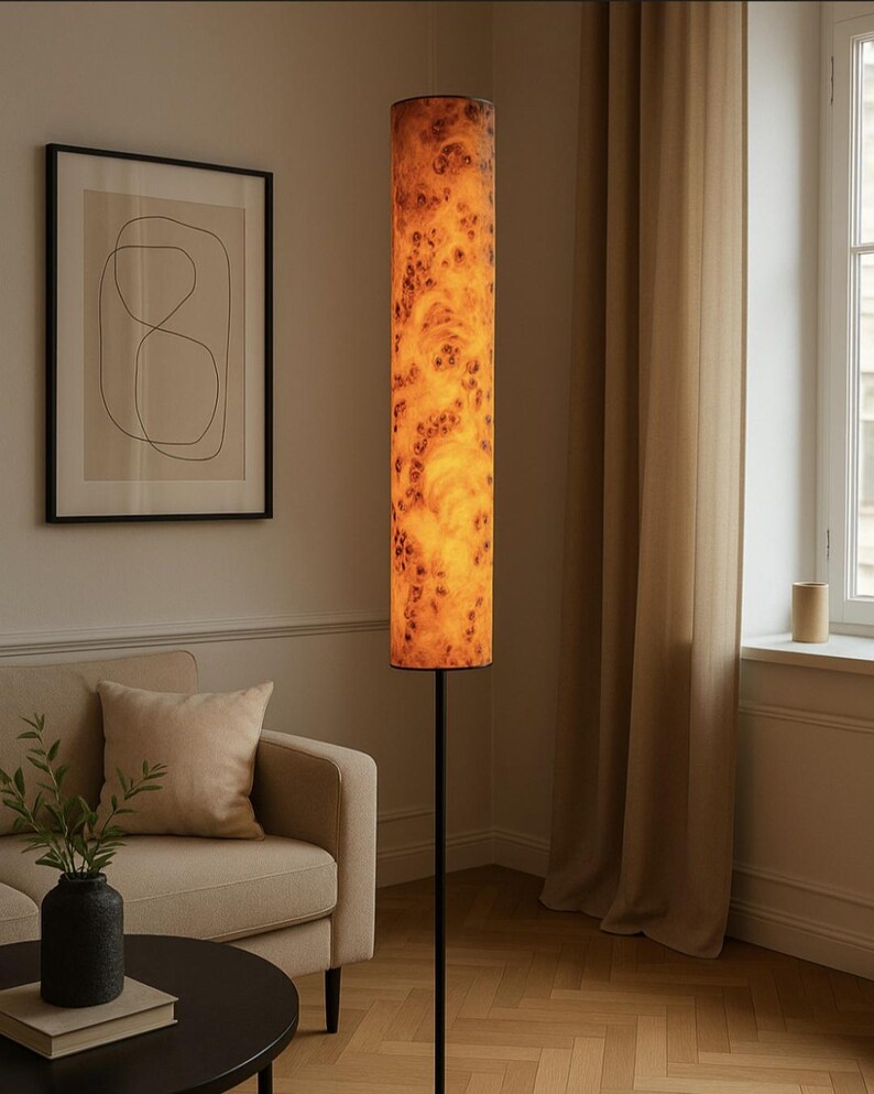 May include: A tall, cylindrical floor lamp with a warm, wood-grain patterned shade. The shade is illuminated, casting a soft glow. The lamp has a slender, black metal stand. The lamp is in a living room setting with a sofa and artwork.