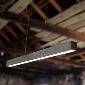 May include: A rectangular wooden pendant light fixture with a bright, linear light source. The light is suspended from the ceiling by thin wires. The wood has a natural grain and a warm, brown tone. The background is a rustic wooden ceiling.