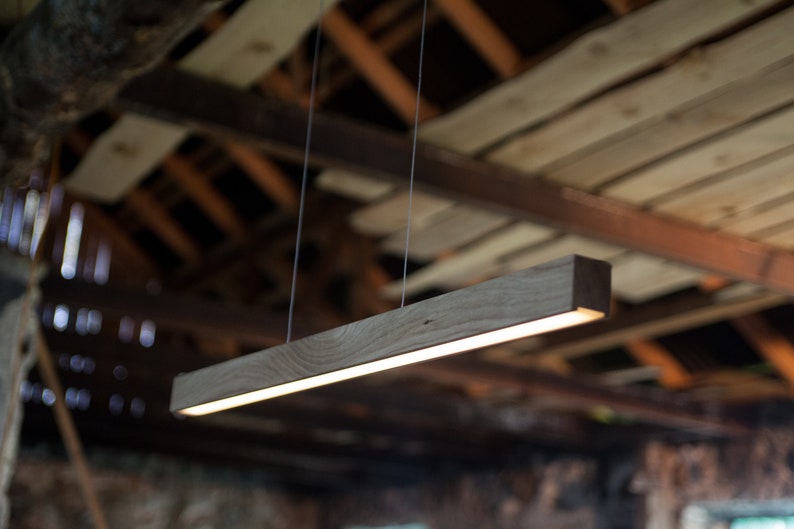 May include: A rectangular wooden pendant light fixture with a bright, linear light source. The light is suspended from the ceiling by thin wires. The wood has a natural grain and a warm, brown tone. The background is a rustic wooden ceiling.