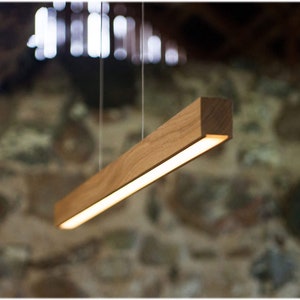May include: A rectangular wooden pendant light fixture. The light is suspended from the ceiling by thin wires. The light has a warm, inviting glow. The wood has a natural grain and color. The background is a blurred stone wall.