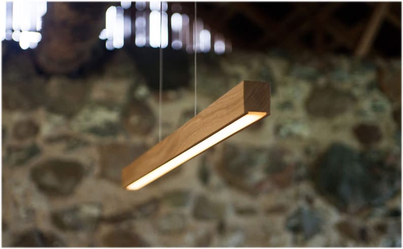May include: A rectangular wooden pendant light fixture. The light is suspended from the ceiling by thin wires. The light has a warm, inviting glow. The wood has a natural grain and color. The background is a blurred stone wall.