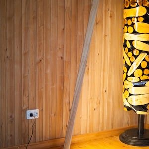 K&ouml;nnte beinhalten: Eine moderne Stehlampe aus Holz mit minimalistischem Design. Die Lampe hat einen hohen, abgewinkelten Holzpfosten und einen runden, beleuchteten Sockel. Die Lampe steht vor einer Holzwand und einem Holzboden.