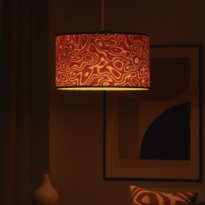 May include: A pendant lamp with a cylindrical lampshade featuring a red and cream abstract pattern. The lamp hangs from the ceiling in a dimly lit room, with a matching patterned pillow on a sofa. A framed artwork and a vase are also visible.