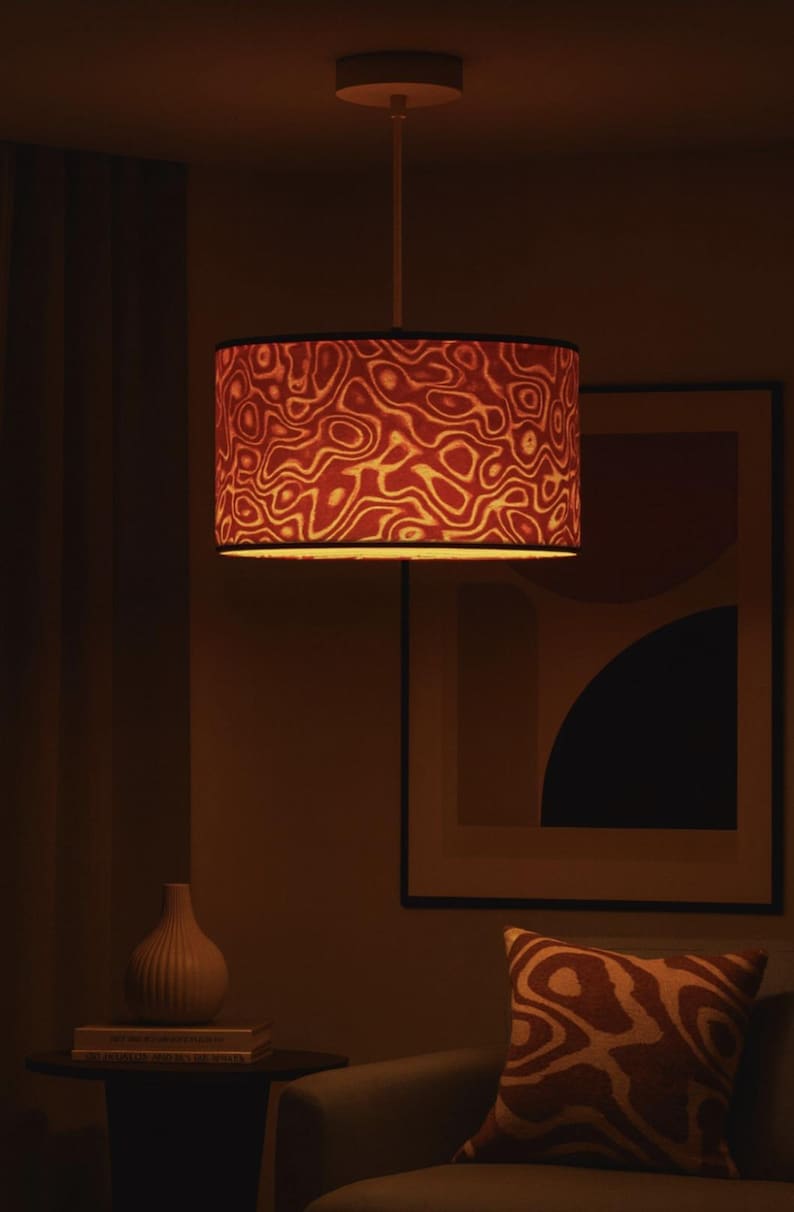May include: A pendant lamp with a cylindrical lampshade featuring a red and cream abstract pattern. The lamp hangs from the ceiling in a dimly lit room, with a matching patterned pillow on a sofa. A framed artwork and a vase are also visible.