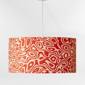 May include: A cylindrical lampshade with a red and cream-colored organic pattern. The lampshade hangs from a white cord and a white ceiling fixture. The pattern resembles wood grain or a topographic map.