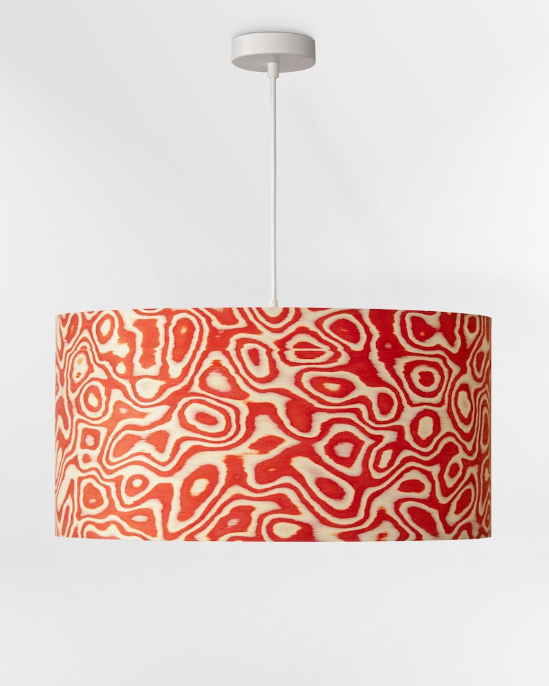May include: A cylindrical lampshade with a red and cream-colored organic pattern. The lampshade hangs from a white cord and a white ceiling fixture. The pattern resembles wood grain or a topographic map.