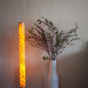 May include: A tall, illuminated floor lamp with a cylindrical shade featuring a warm, wood-grain pattern. The lamp stands on a circular metal base. Beside it, a white textured vase holds branches with green leaves and pink flowers.