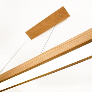 May include: A modern wooden light fixture with a rectangular design. The fixture has a light-colored wood finish and a bright, white light panel. The fixture is suspended from the ceiling by thin metal wires. The overall design is minimalist and contemporary.