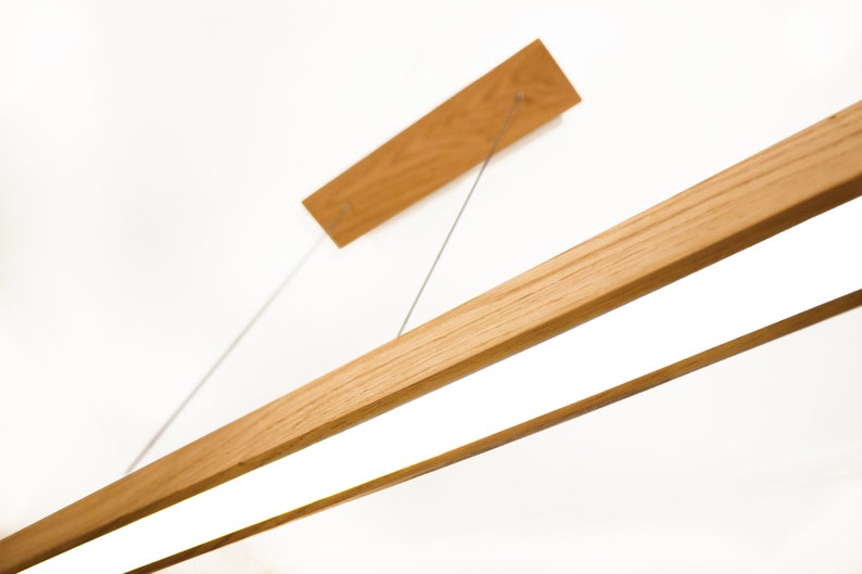 May include: A modern wooden light fixture with a rectangular design. The fixture has a light-colored wood finish and a bright, white light panel. The fixture is suspended from the ceiling by thin metal wires. The overall design is minimalist and contemporary.