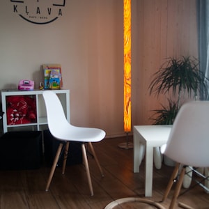 May include: Interior shot of a modern living space with a tall, illuminated floor lamp. The lamp has a vibrant, swirling orange and yellow pattern. White chairs with wooden legs and a small white table are also visible.
