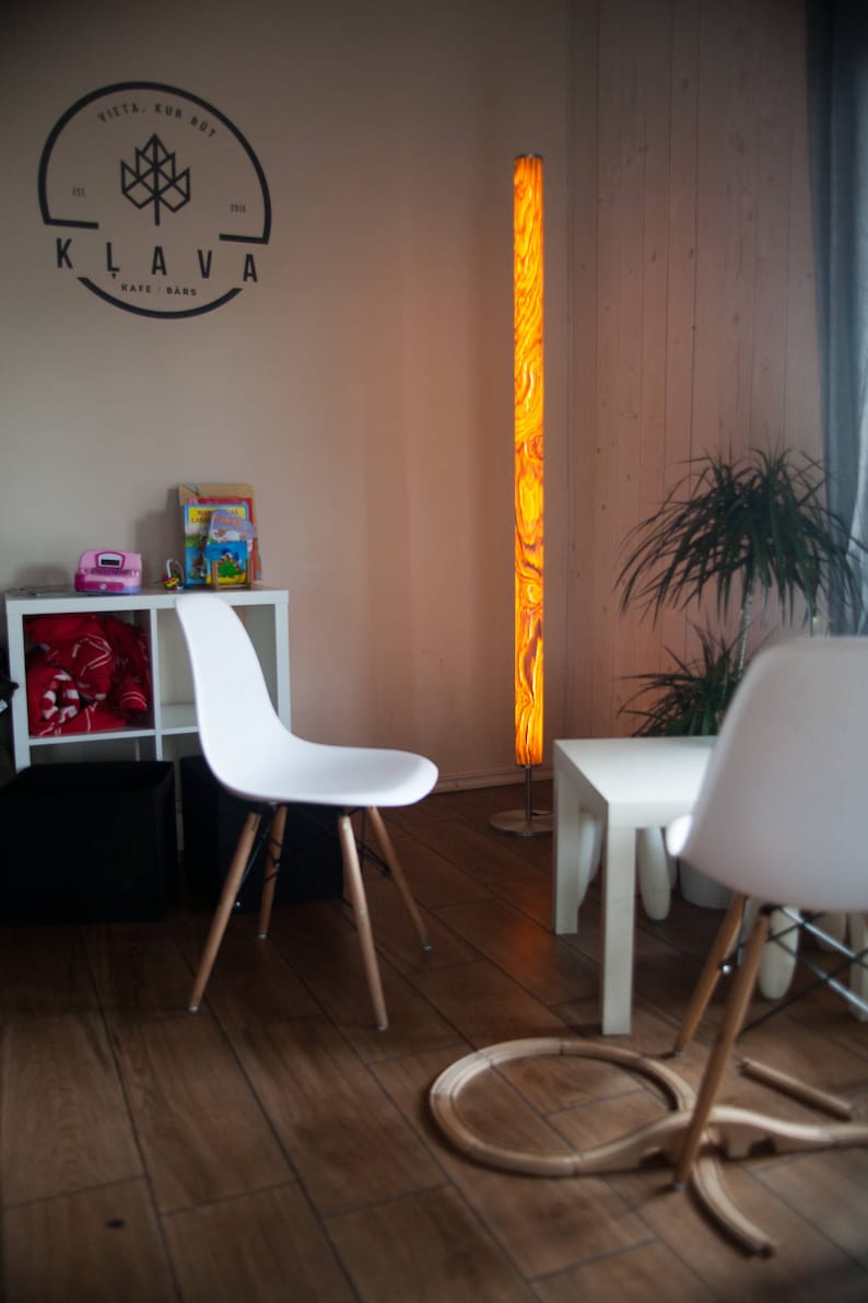 May include: Interior shot of a modern living space with a tall, illuminated floor lamp. The lamp has a vibrant, swirling orange and yellow pattern. White chairs with wooden legs and a small white table are also visible.
