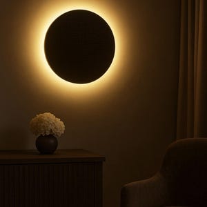 Puede incluir: Una moderna lámpara de pared circular con un cálido efecto de halo brillante. El disco oscuro y texturizado está montado en una pared, con una luz ambiental suave que lo rodea. Un mueble de madera oscura y un jarrón con flores blancas están en primer plano.