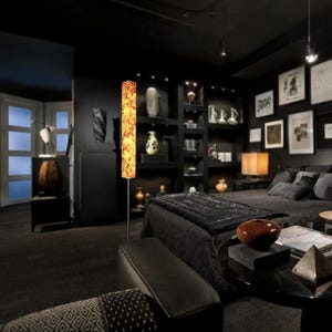 May include: A modern bedroom with black walls, furniture, and decor. A tall, decorative floor lamp with a warm, patterned shade stands out. The room includes a bed, shelves with decor, and framed artwork.