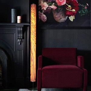 May include: A tall, cylindrical floor lamp with a wood grain pattern stands next to a burgundy armchair. The lamp's warm, amber glow contrasts with the dark walls and fireplace. A large floral painting hangs above the chair, adding a pop of color to the room.