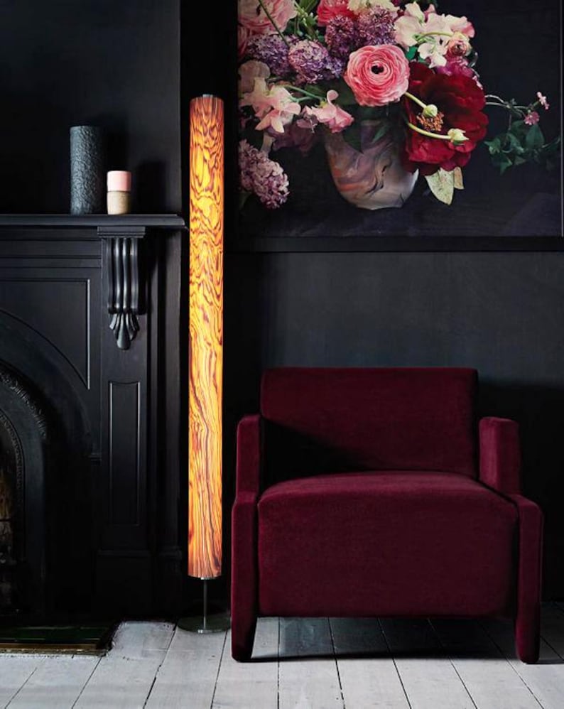 May include: A tall, cylindrical floor lamp with a wood grain pattern stands next to a burgundy armchair. The lamp's warm, amber glow contrasts with the dark walls and fireplace. A large floral painting hangs above the chair, adding a pop of color to the room.