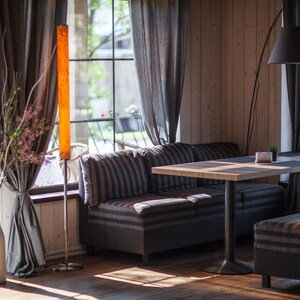 May include: Interior shot of a restaurant or cafe with natural light. Features a striped sofa, wooden table, and a tall, illuminated floor lamp. Gray curtains frame large windows overlooking greenery. A vase with branches and flowers adds a decorative touch.