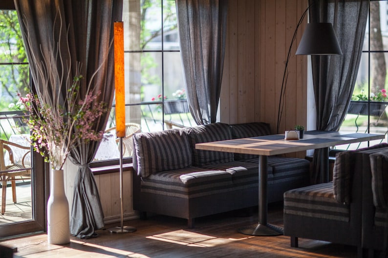 May include: Interior shot of a restaurant or cafe with natural light. Features a striped sofa, wooden table, and a tall, illuminated floor lamp. Gray curtains frame large windows overlooking greenery. A vase with branches and flowers adds a decorative touch.