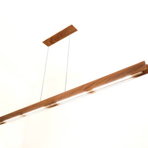 May include: A contemporary wooden pendant light. The light fitting features a rectangular wooden frame with integrated LED lighting. It is suspended from the ceiling by thin wires, creating a minimalist design. The light measures approximately 120 cm in length.