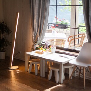 K&ouml;nnte beinhalten: Ein modernes Interieur mit einem wei&szlig;en Tisch und Hockern, einer hohen Holz-Stehleuchte und einem wei&szlig;en Stuhl. Ein gro&szlig;es Fenster mit grauen Vorh&auml;ngen blickt auf eine Au&szlig;enterrasse mit St&uuml;hlen und Pflanzen. Warme, einladende Atmosph&auml;re.