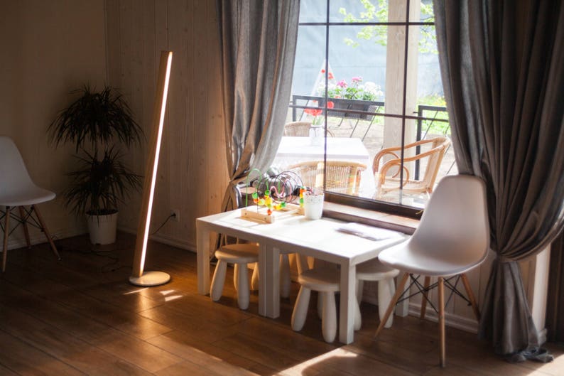 K&ouml;nnte beinhalten: Ein modernes Interieur mit einem wei&szlig;en Tisch und Hockern, einer hohen Holz-Stehleuchte und einem wei&szlig;en Stuhl. Ein gro&szlig;es Fenster mit grauen Vorh&auml;ngen blickt auf eine Au&szlig;enterrasse mit St&uuml;hlen und Pflanzen. Warme, einladende Atmosph&auml;re.