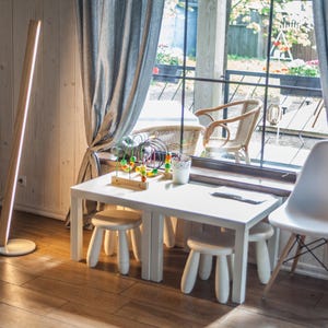 K&ouml;nnte beinhalten: Ein modernes Interieur mit einem wei&szlig;en Tisch und Hockern, einem wei&szlig;en Stuhl und einer Holzstehleuchte mit vertikalem Lichtstreifen. Der Raum hat ein Fenster mit grauen Vorh&auml;ngen und Blick auf eine Au&szlig;enterrasse.