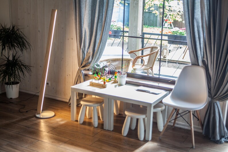 K&ouml;nnte beinhalten: Ein modernes Interieur mit einem wei&szlig;en Tisch und Hockern, einem wei&szlig;en Stuhl und einer Holzstehleuchte mit vertikalem Lichtstreifen. Der Raum hat ein Fenster mit grauen Vorh&auml;ngen und Blick auf eine Au&szlig;enterrasse.