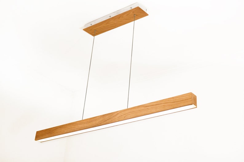 May include: A modern, rectangular pendant light fixture. The light features a long, wooden beam with a bright, white light strip. The fixture is suspended by thin, silver cables from a rectangular wooden ceiling mount.